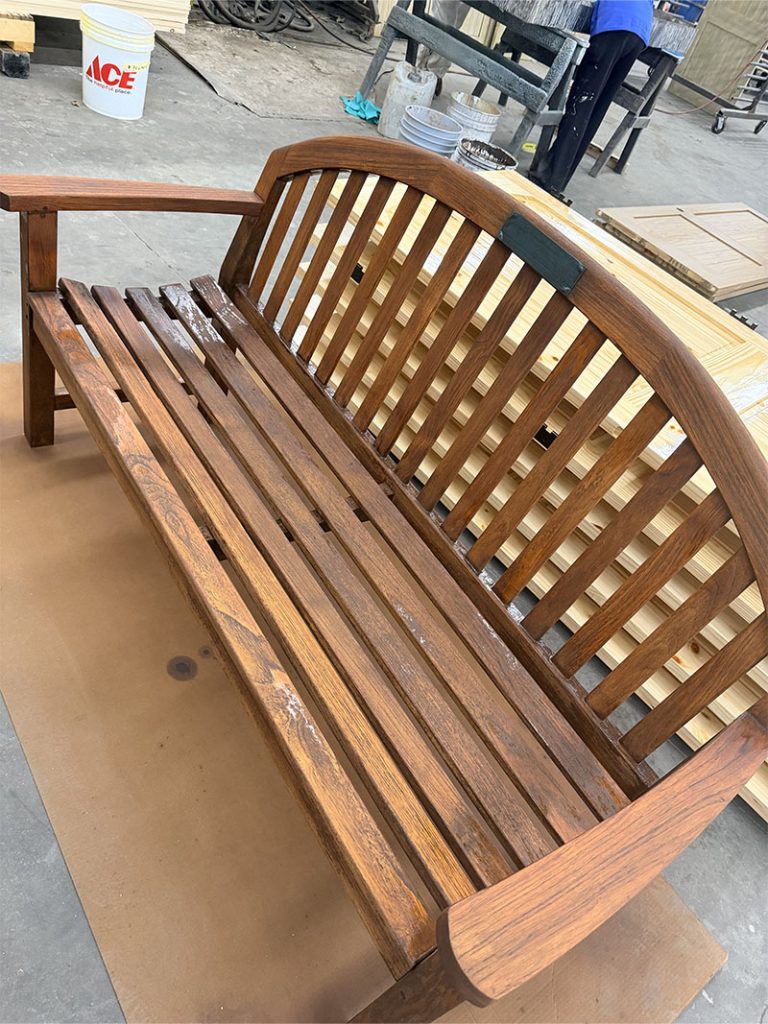 A wooden slatted bench with a curved backrest sits on a cardboard sheet in a workshop, surrounded by wooden planks and tools.