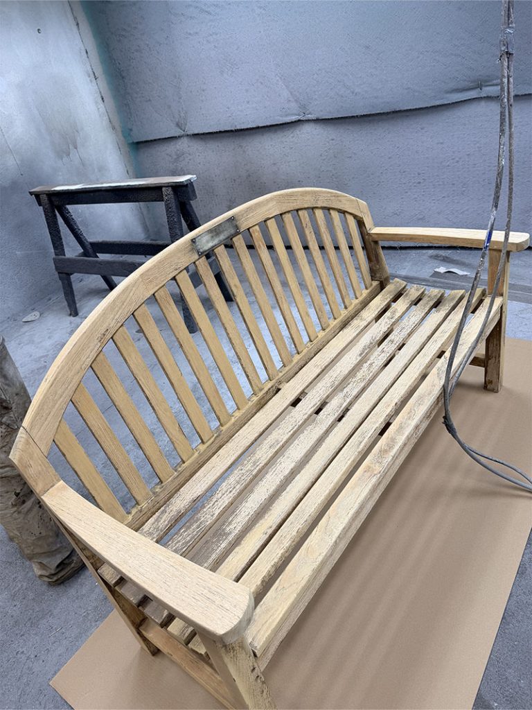 A weathered wooden bench with vertical slats sits on a piece of cardboard in a workshop. Another small, dark-colored table is visible in the background. The area appears to be set up for restoration or refinishing work.