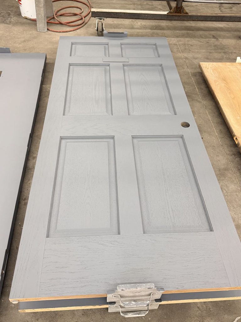 A gray, six-panel door lies flat on a workshop floor with a hole for a doorknob. Tools, a coiled orange cable, and another door are visible in the background.