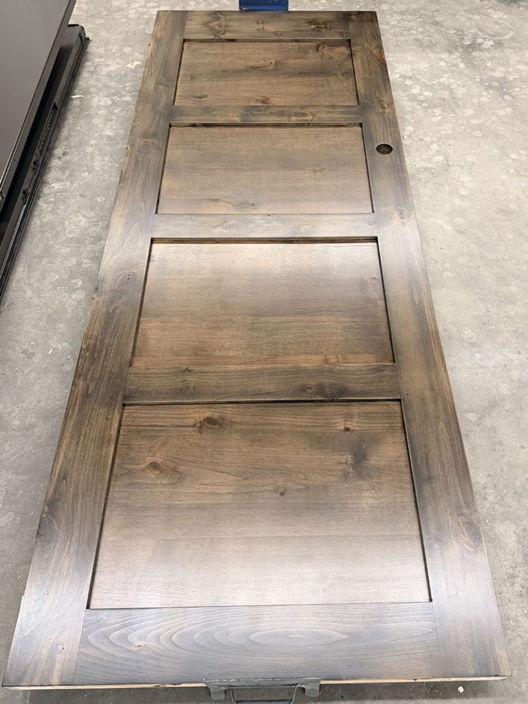 A wooden door with three rectangular panels lying flat on a concrete floor. The door has a natural wood grain finish and a hole for a doorknob on the right side.