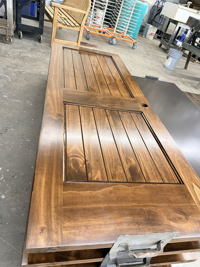 A freshly stained wooden door with a rich brown finish lies flat on a workbench in a workshop, surrounded by tools, equipment, and furniture in the background.