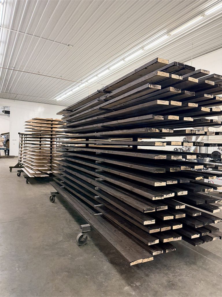 Stacks of long, dark wooden planks are arranged on metal racks with wheels in a spacious, well-lit industrial workshop with a high ceiling.
