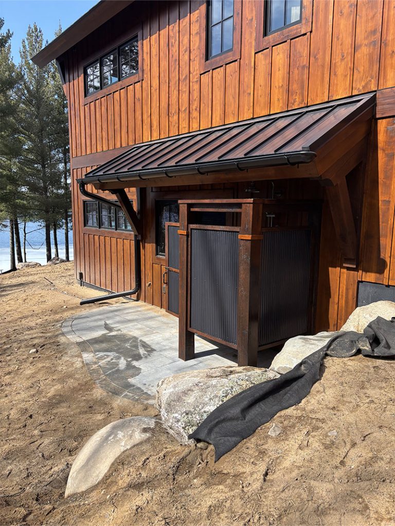 A wooden house with dark trim and a metal roof extension over a small porch area, near trees and a lake. Large rocks and exposed soil are in the foreground with some landscaping fabric.