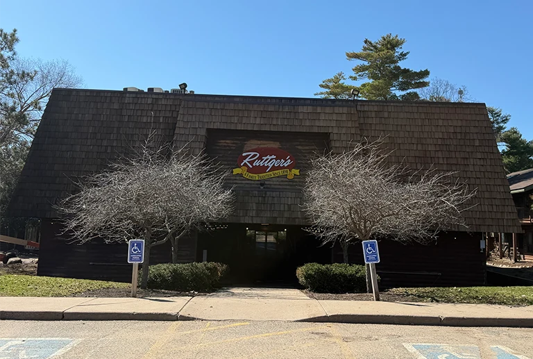 A brown, wood-paneled building with a sign that reads "Rutgers" above the entrance, flanked by two leafless trees and two accessible parking signs in front, on a clear, sunny day.