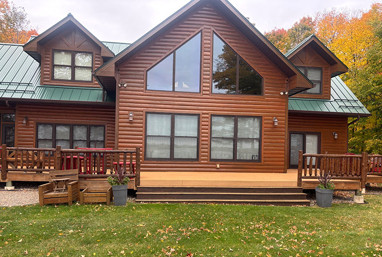 A large wooden cabin with a green metal roof, large triangular windows, two porches, and potted plants sits in front of a grassy lawn with autumn trees in the background.