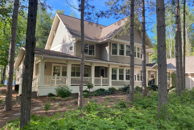A two-story beige house with white trim and a large wraparound porch, surrounded by tall trees and greenery under a blue sky.