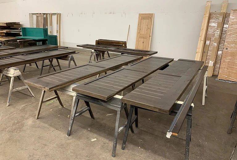 Several wooden doors and panels are laid out on sawhorses in a workshop, with additional wood pieces, doors, and materials stacked against the walls in the background.