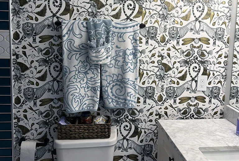A bathroom features bold wallpaper with animal and leaf patterns, a white toilet with a basket on top, blue and white patterned towels hanging above, and part of a marble countertop and sink on the right.